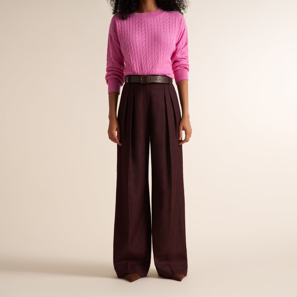 A model is shown from the neck down wearing a bright pink cable-knit sweater and dark brown wide-leg pleated trousers with a thin dark belt, set against a plain beige studio background.