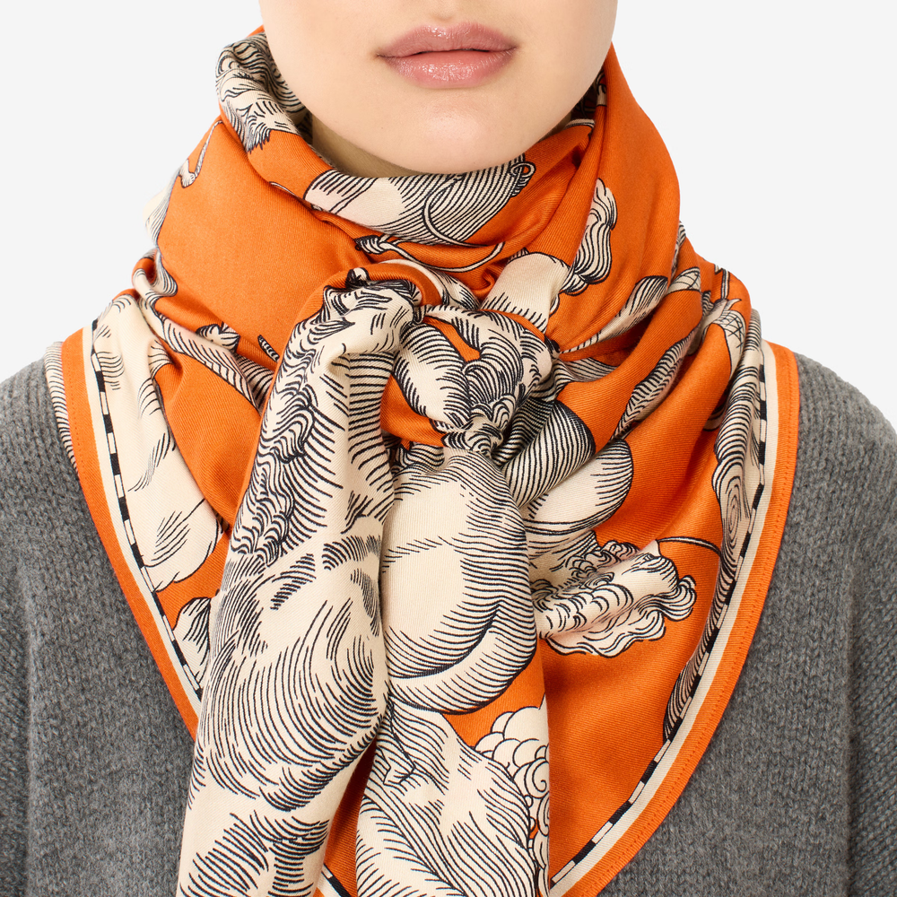 An orange silk scarf with a black and cream classical-style illustration print, styled around the neck of a person wearing a gray knit sweater against a light gray background.