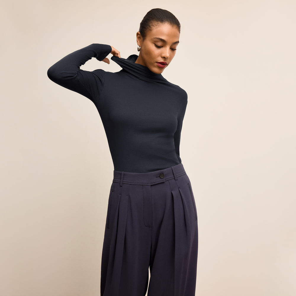 Woman wearing a navy turtleneck and dark pants against a beige background