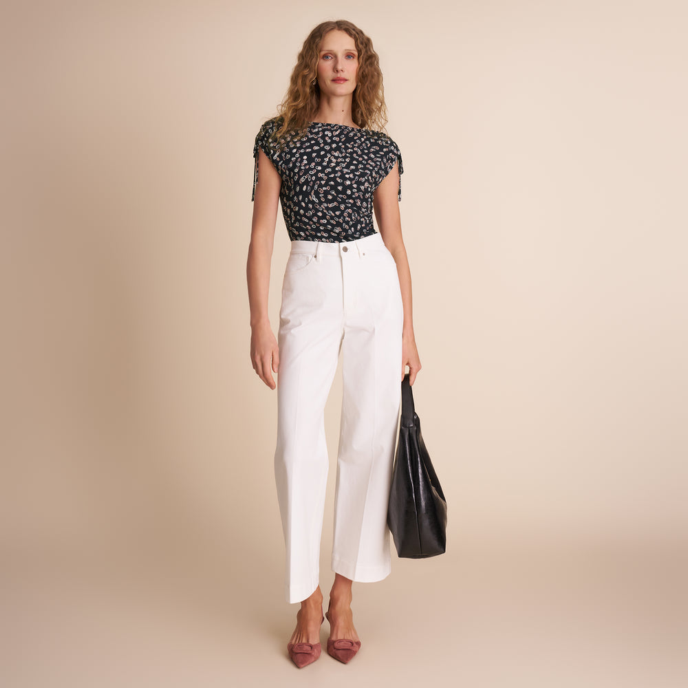 Woman wearing a black floral top and white pants, holding a black bag on a beige background