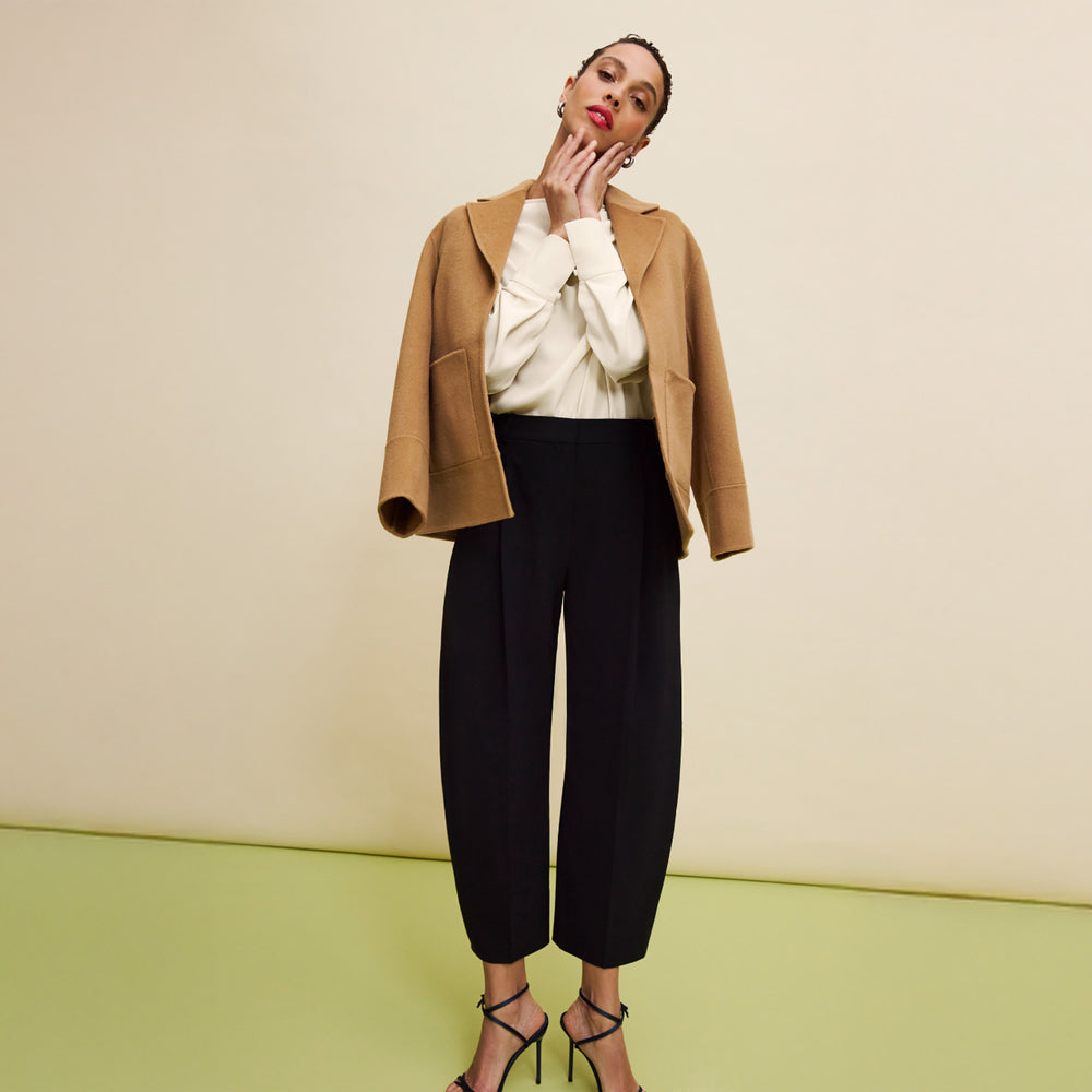 Woman wearing a tan coat, white sweater, and black pants on a beige and green background
