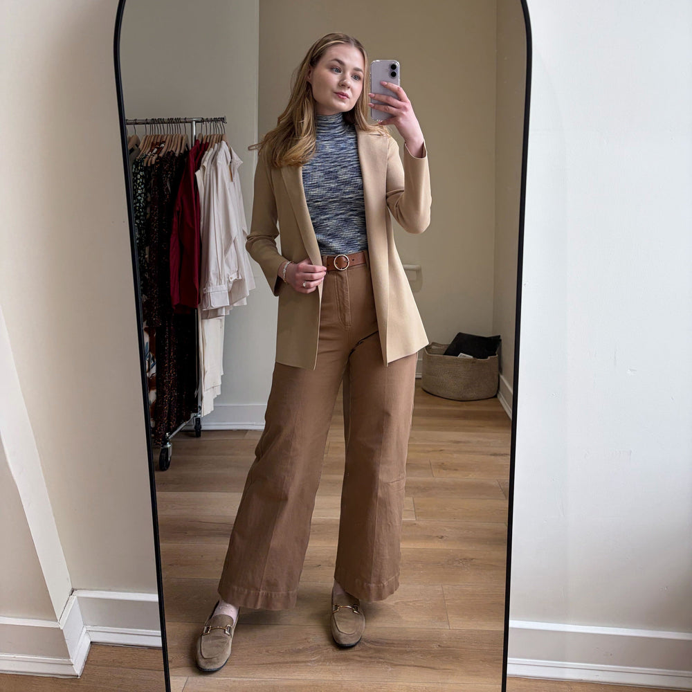 Person taking a mirror selfie wearing a beige blazer and pants in a room with a clothing rack.