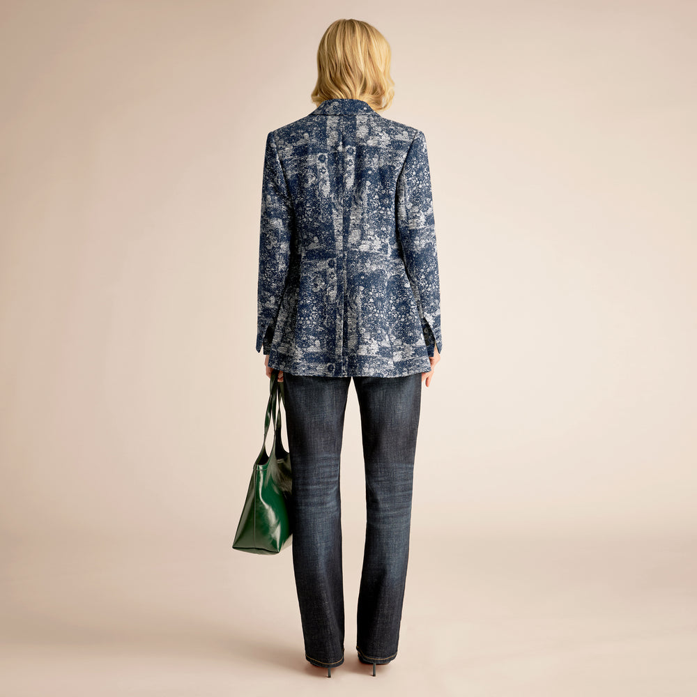 Person wearing a blue patterned jacket and dark jeans, holding a green bag on a beige background