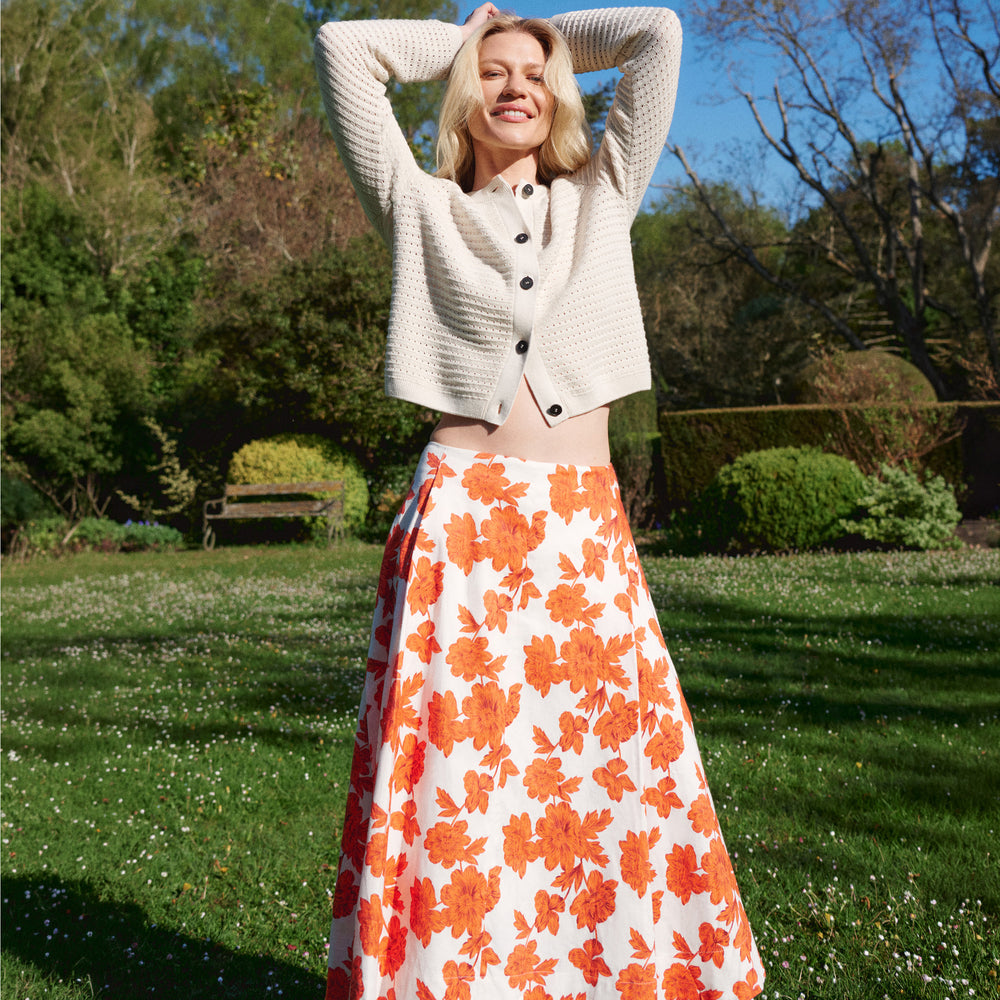 Woman in a white cardigan and orange floral skirt standing in a park.