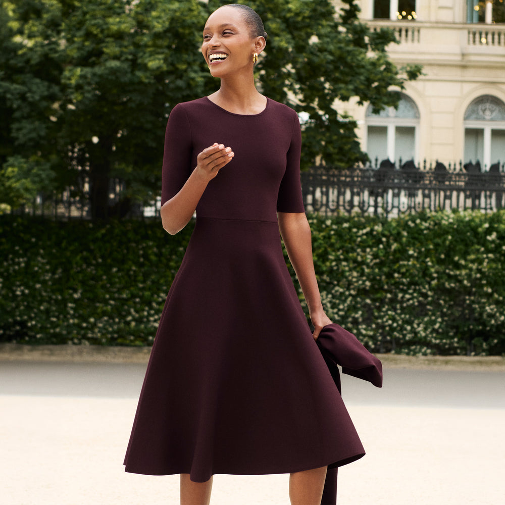 A woman wearing a dark burgundy midi dress with short sleeves and a flared skirt, holding a matching clutch bag, standing outdoors in front of a green hedge and a building.