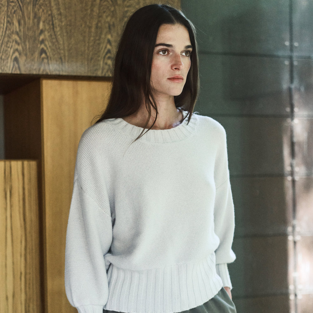 Woman wearing a light gray sweater and green pants standing against a wooden wall.