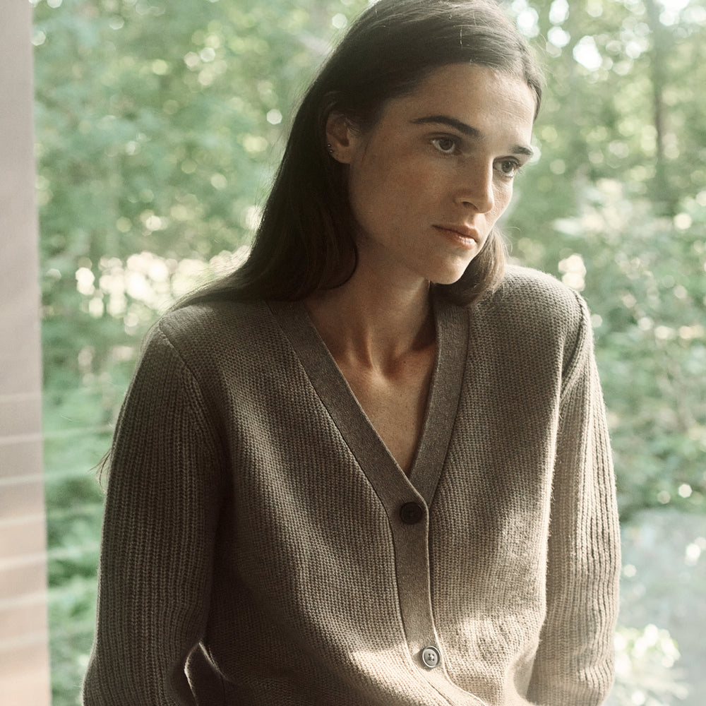 Woman wearing a brown cardigan and white shorts sitting by a window with greenery outside.