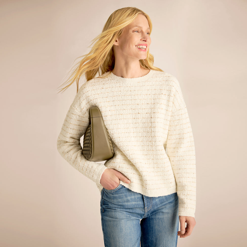 Woman wearing a cream sweater and blue jeans, holding a beige handbag against a plain background
