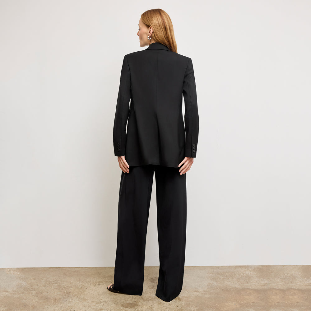 Person wearing a black suit standing against a plain white wall.