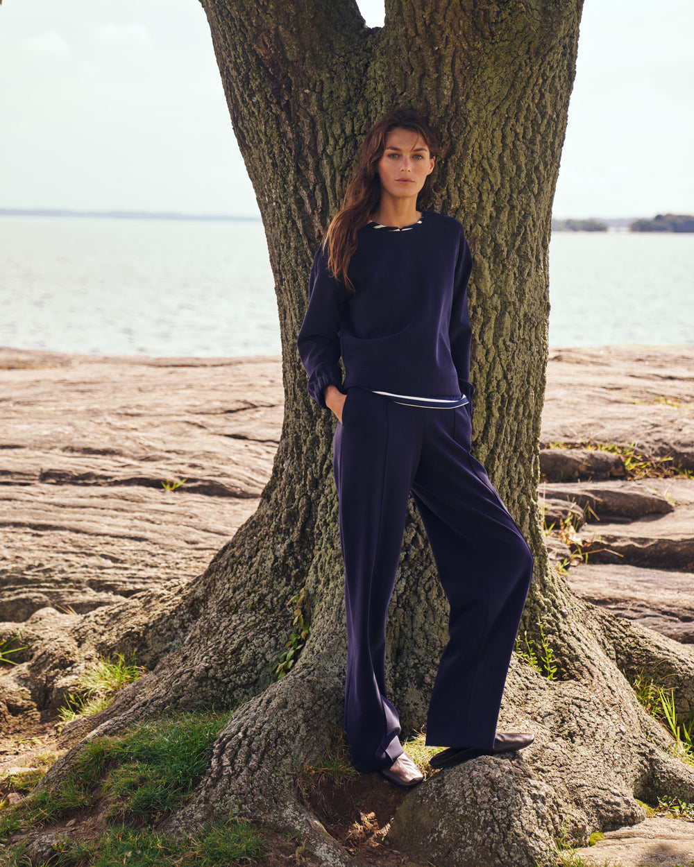 A woman wearing a navy blue two-piece lounge set with a white stripe detail, leaning against a large tree trunk on a rocky shoreline with water in the background.