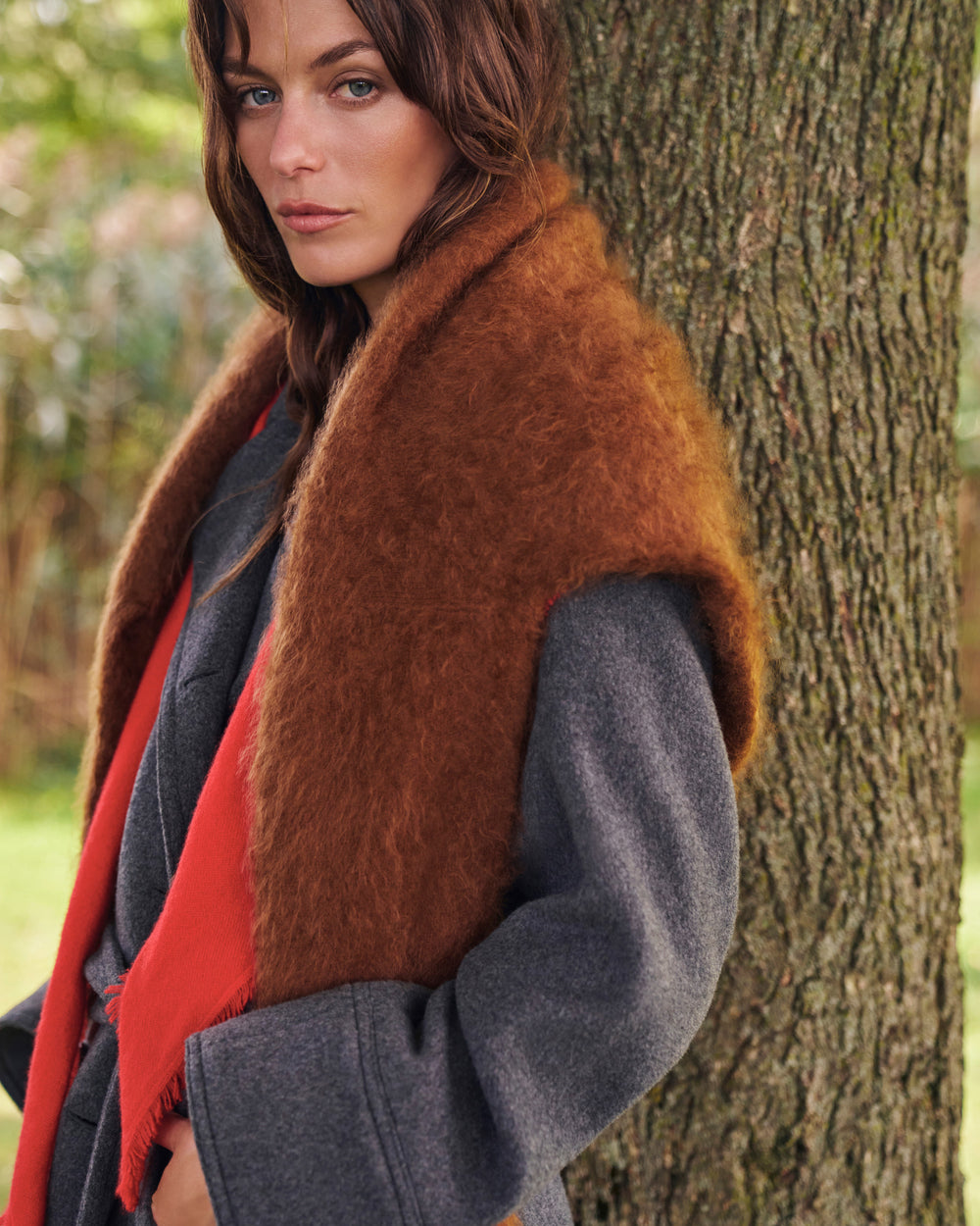 A woman outdoors leaning against a tree, wearing a brown shaggy faux-fur collar or wrap over a gray coat and a bright red scarf.
