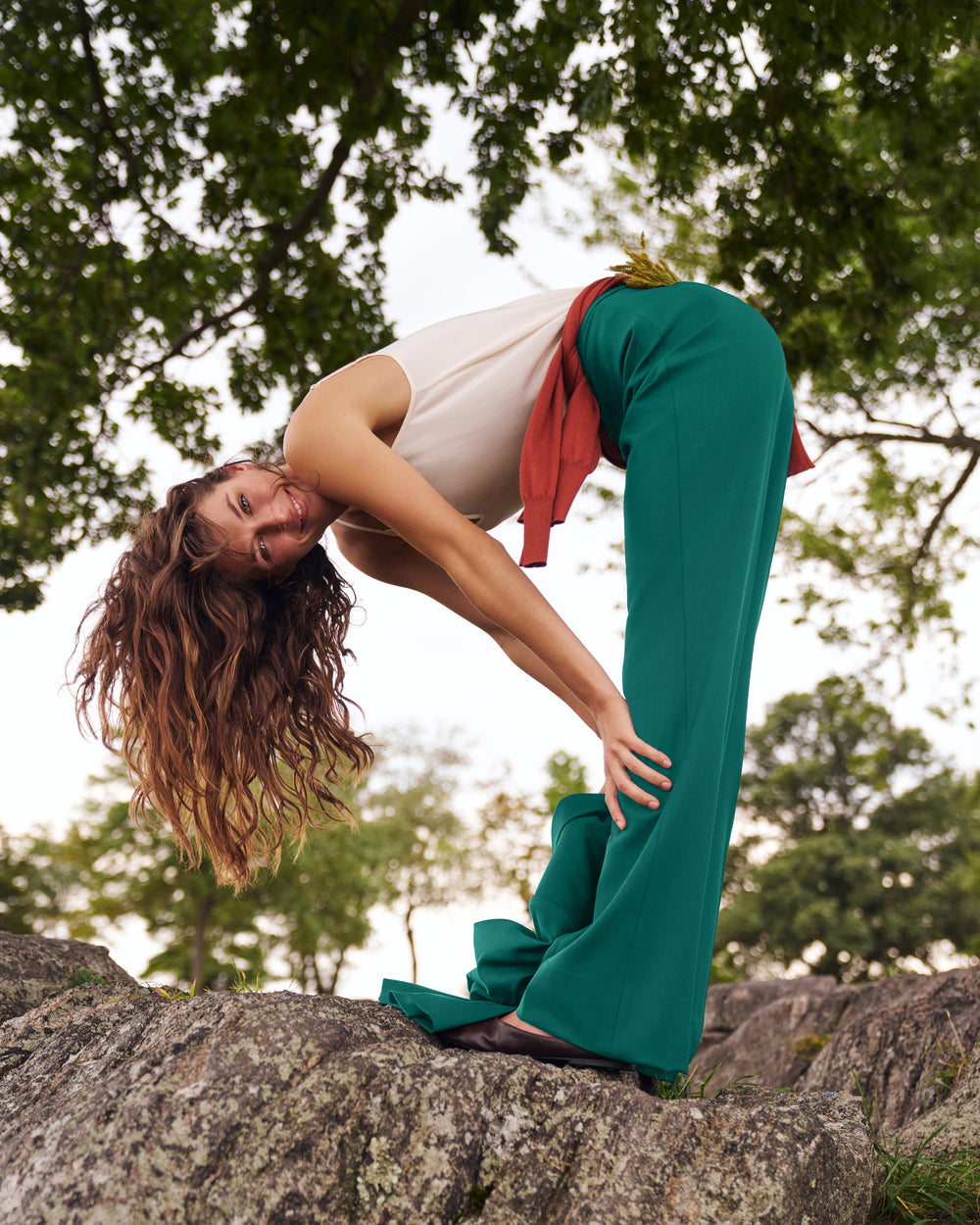 A woman in a yoga pose outdoors on a large rock, wearing teal wide-leg pants, a cream tank top, and a red shirt tied around her waist, with trees in the background.