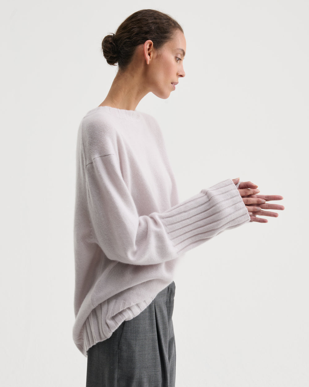 A woman in profile wearing a pale blush pink knit sweater with ribbed long sleeves and dark gray trousers, photographed against a clean off-white studio background.