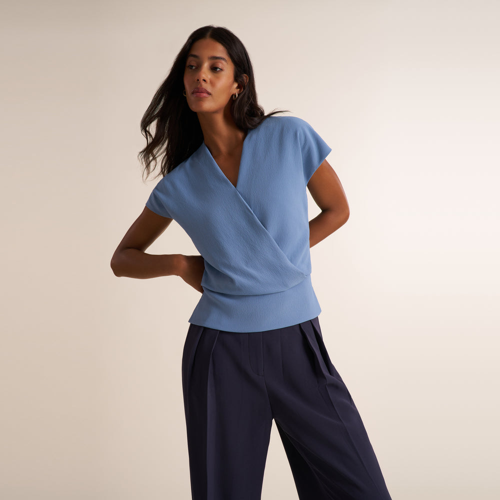 A woman modeling a light blue wrap-style short-sleeve top with dark navy wide-leg trousers, posed against a solid warm beige studio background.