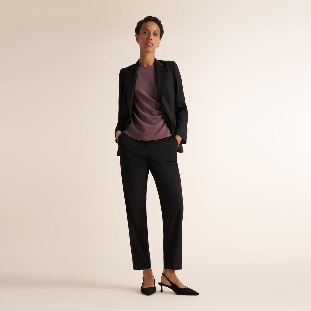 A model stands in a studio wearing a black tailored blazer and matching black trousers over a mauve-purple top, with black pointed heels, against a plain warm off-white background.