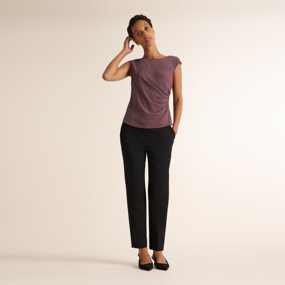 A woman modeling a mauve-purple short-sleeve top and black trousers against a plain beige studio background.