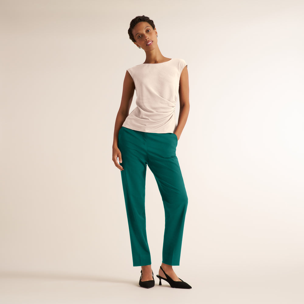 A model wearing a sleeveless cream top with a gathered side detail and teal green trousers, paired with black pointed-toe heels, standing against a plain light beige studio background.