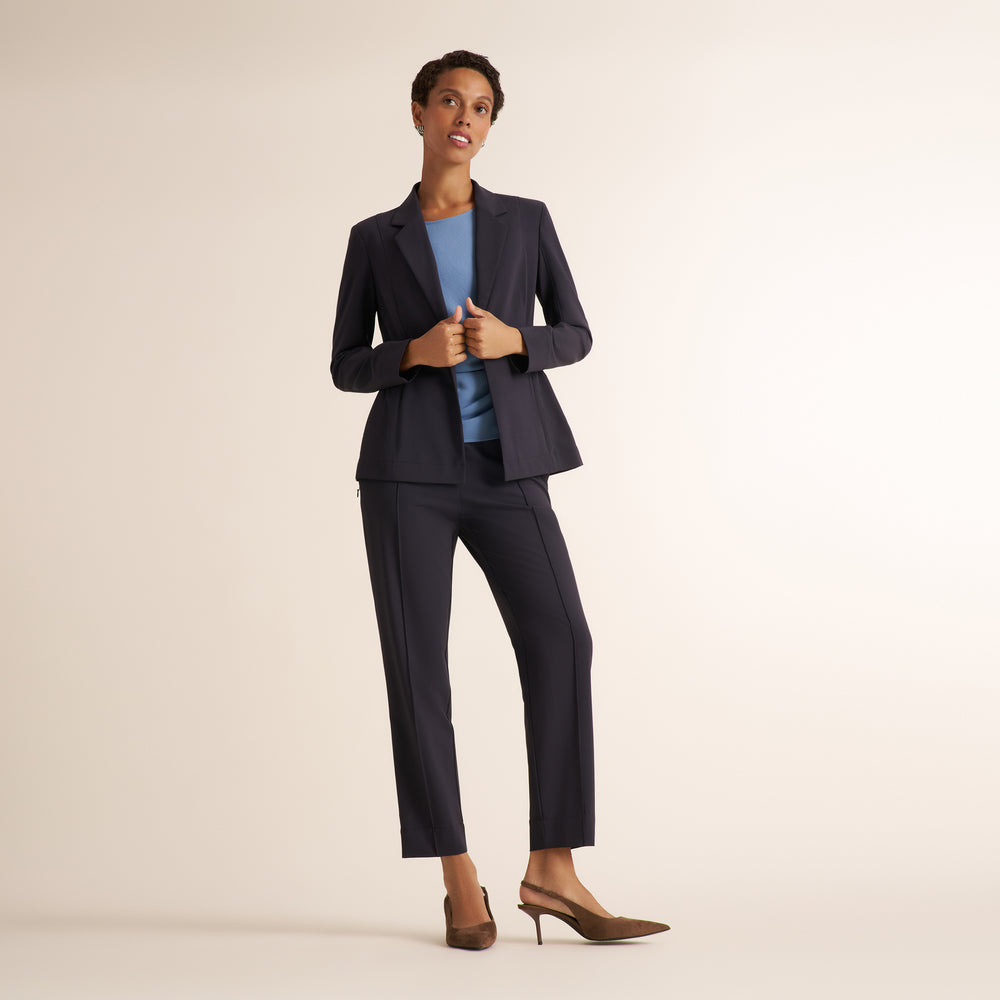 A woman modeling a matching charcoal pantsuit with a blazer and straight-leg trousers, styled with a light blue top and brown strappy heels against a plain beige studio