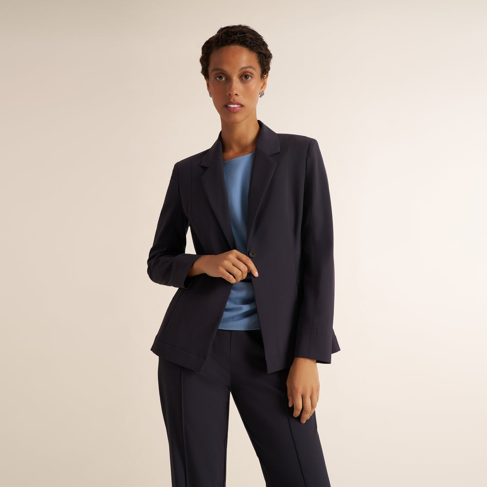 A woman modeling a matching charcoal pantsuit with a blazer and straight-leg trousers, styled with a light blue top and brown strappy heels against a plain beige studio