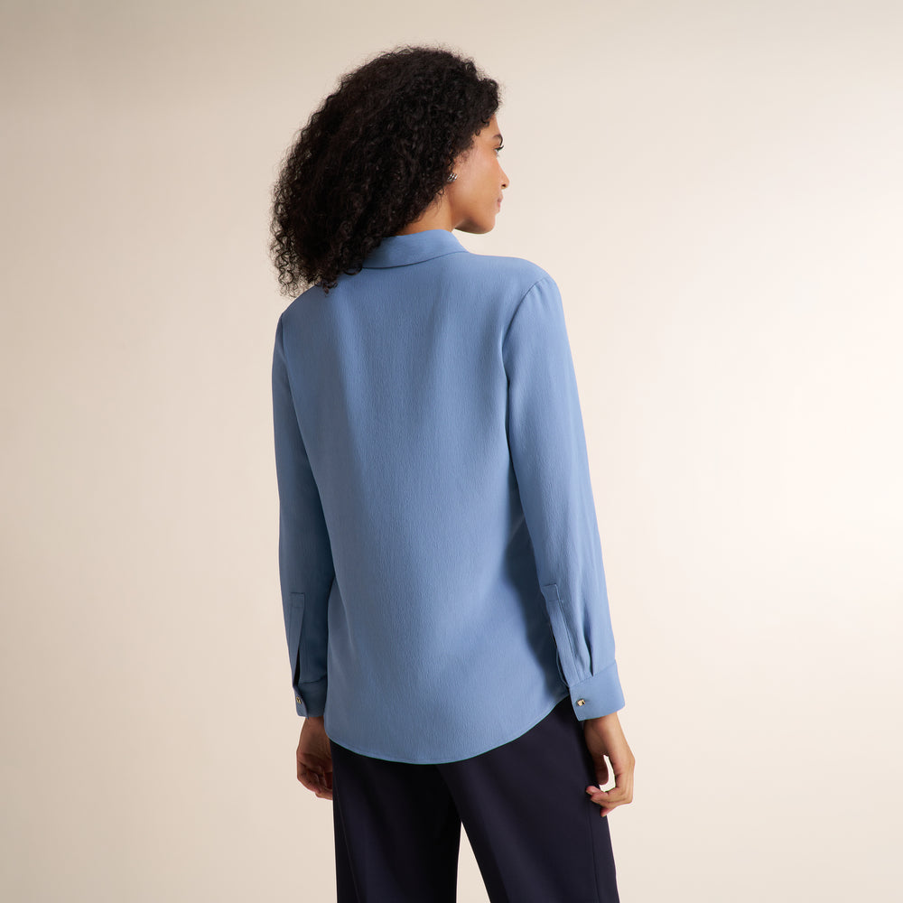 A woman with curly dark hair is shown from the back wearing a light blue long-sleeve blouse and dark navy trousers, standing against a plain warm beige studio background.