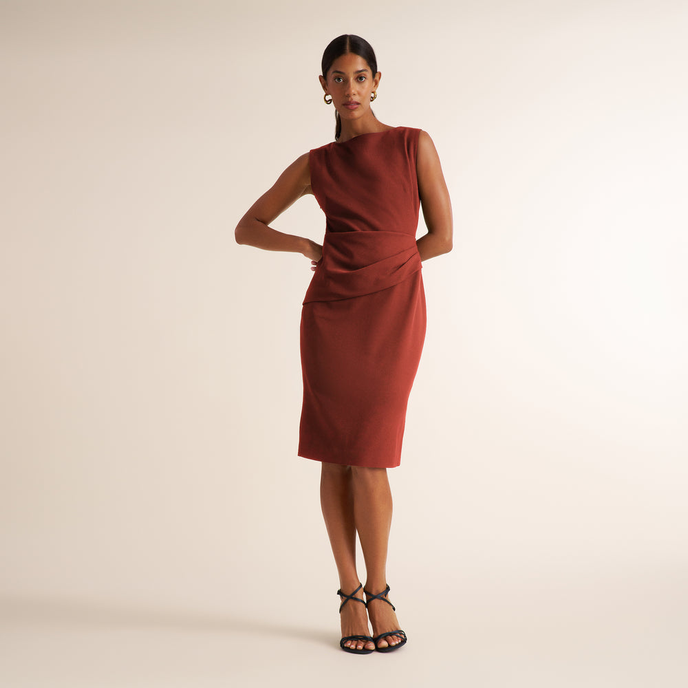 A woman modeling a sleeveless, knee-length terracotta dress with a draped waist detail, paired with black strappy heels, against a solid light beige studio background.