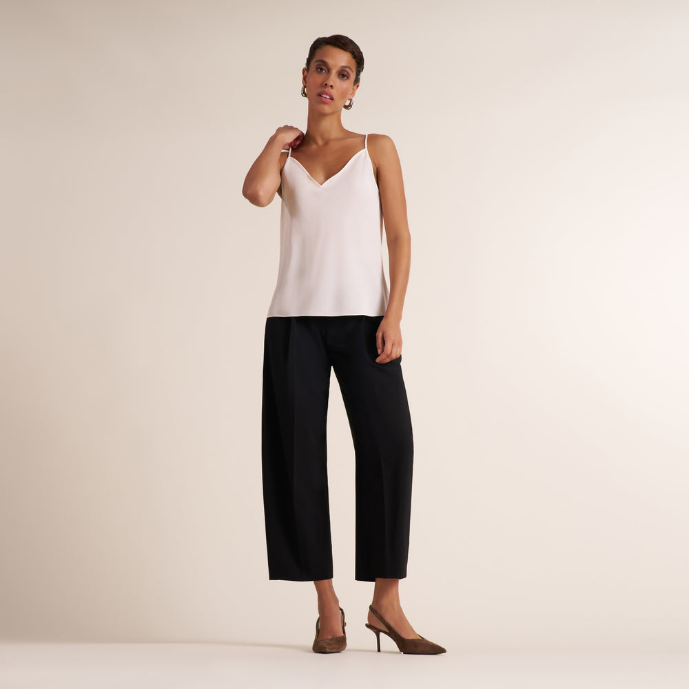 A model wearing a white spaghetti-strap camisole top and black cropped wide-leg trousers, paired with brown pointed-toe mule heels, standing against a plain beige studio background.