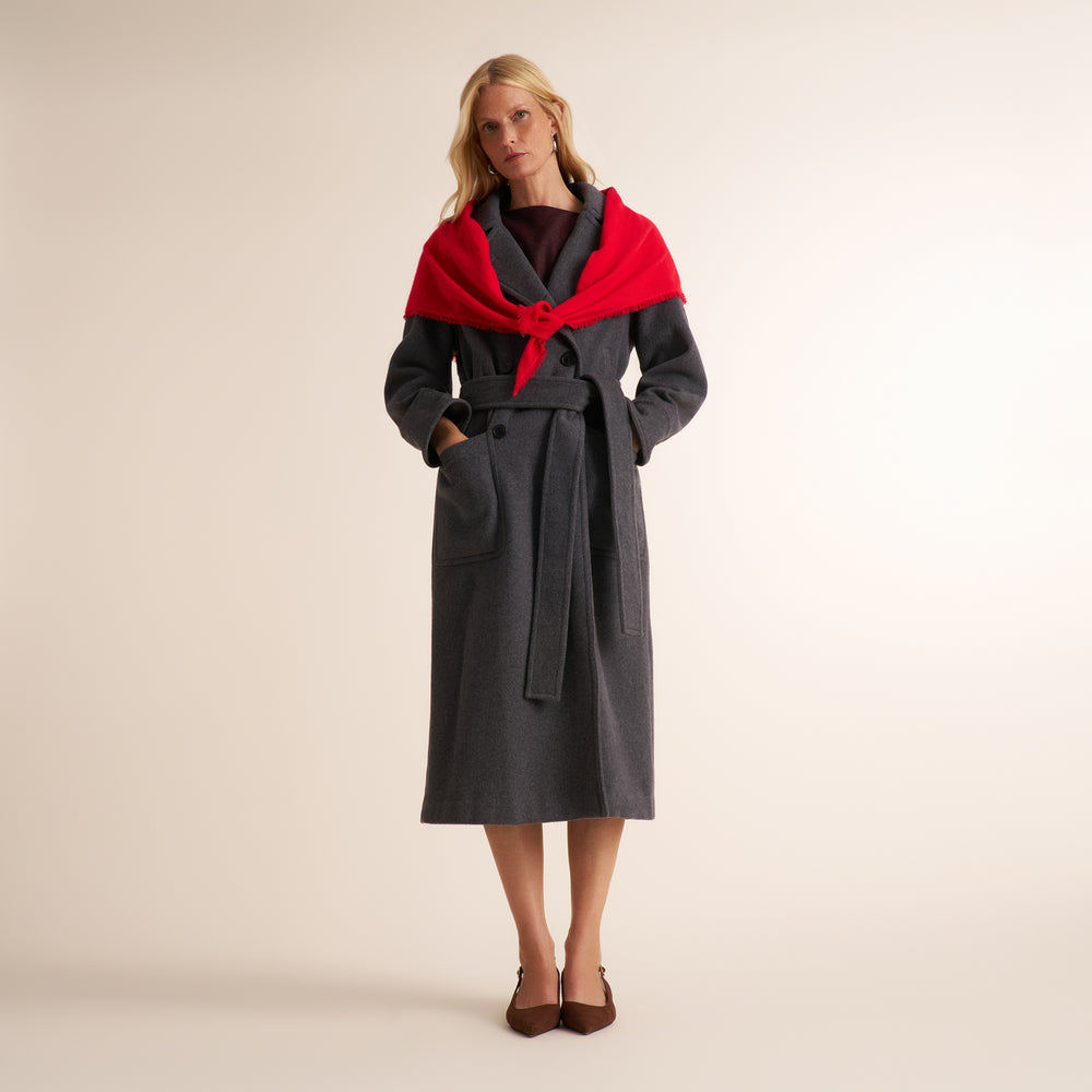 A woman modeling a dark gray wool coat with a bright red shawl wrapped around her shoulders, set against a plain beige studio background.
