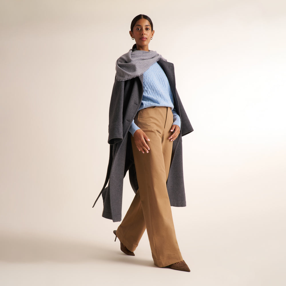 A model is shown wearing a stylish outfit consisting of a light blue ribbed knit sweater, wide-leg tan trousers, a gray wrap scarf, and a long dark gray open-front coat, set against a solid warm beige studio background.