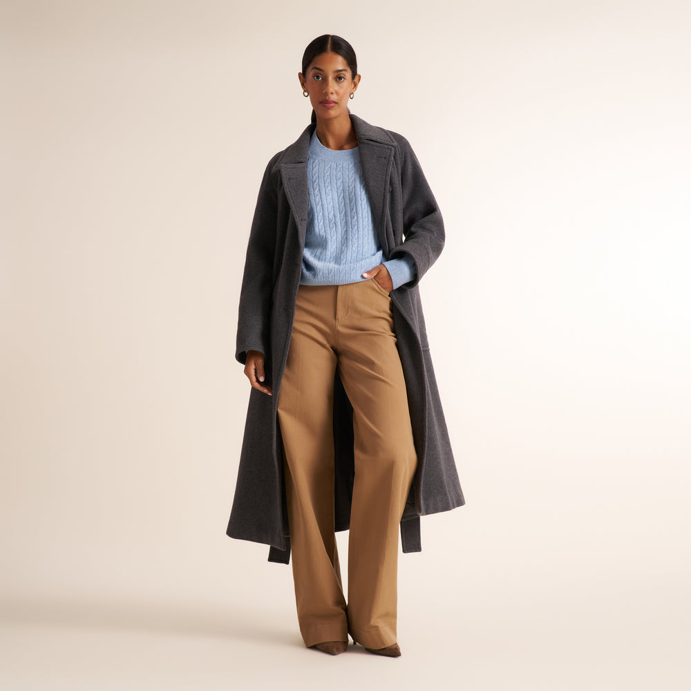 A model stands against a warm beige studio background wearing a long charcoal gray coat over a light blue cable-knit sweater and tan wide-leg trousers.