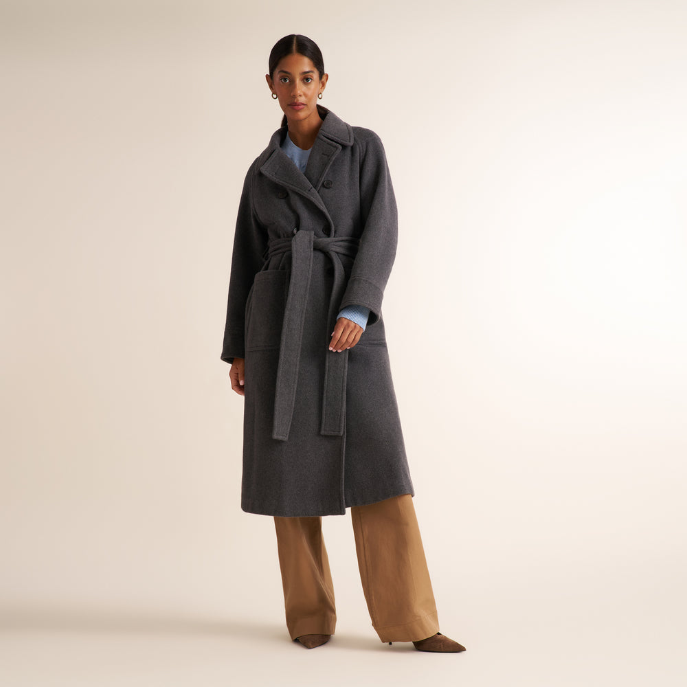 A model stands against a warm beige studio background wearing a dark gray belted coat over a light blue top and wide-leg tan pants.