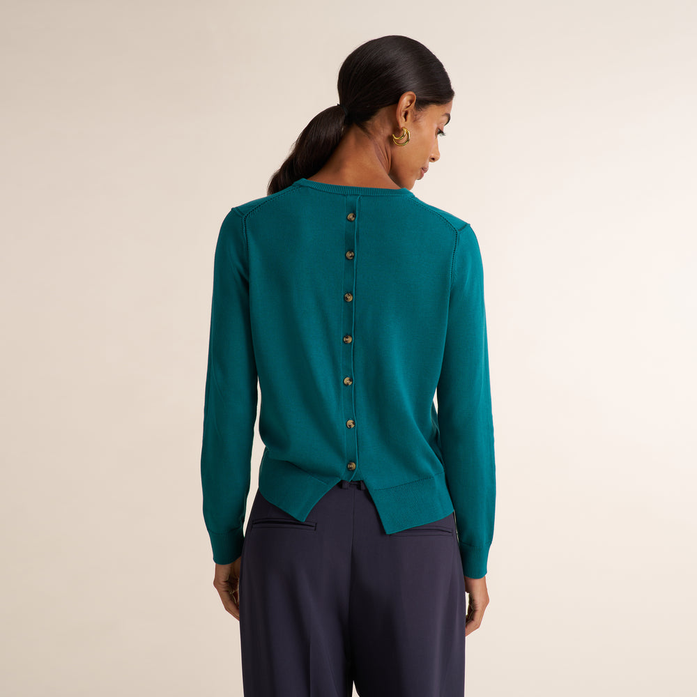 A woman shown from the back wearing a teal long-sleeve knit top with a vertical row of gold buttons down the center, paired with dark navy pants, against a plain light beige studio background.