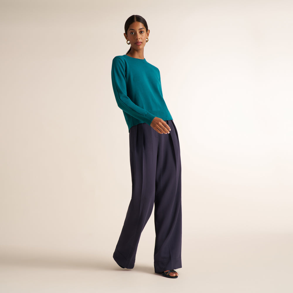 A model stands in a studio wearing a teal long-sleeve knit top and dark navy wide-leg trousers, paired with black sandals against a warm off-white background.