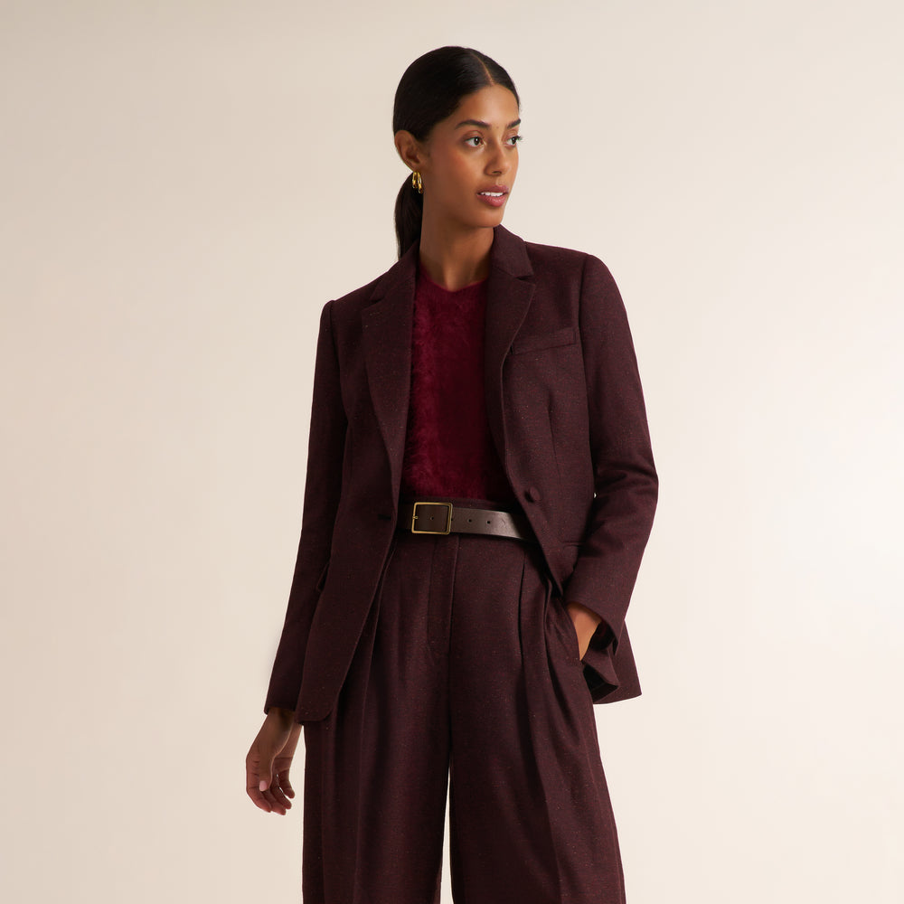 A woman modeling a tailored dark burgundy blazer with matching high-waisted trousers, worn over a textured burgundy top and a dark belt, against a plain light beige studio background.