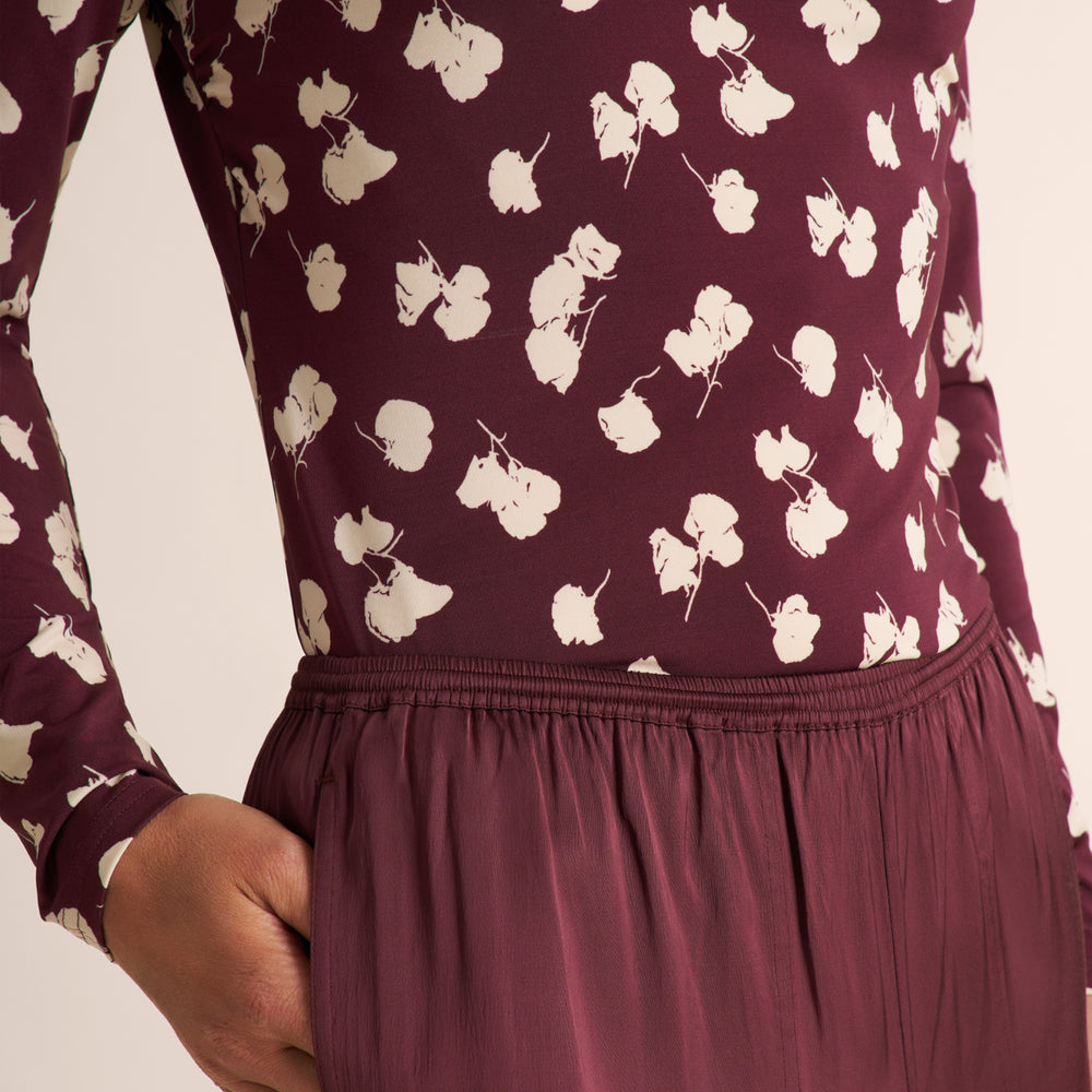 Close-up of a person wearing a long-sleeve burgundy top with a white floral print and a matching pleated skirt, against a light beige background.