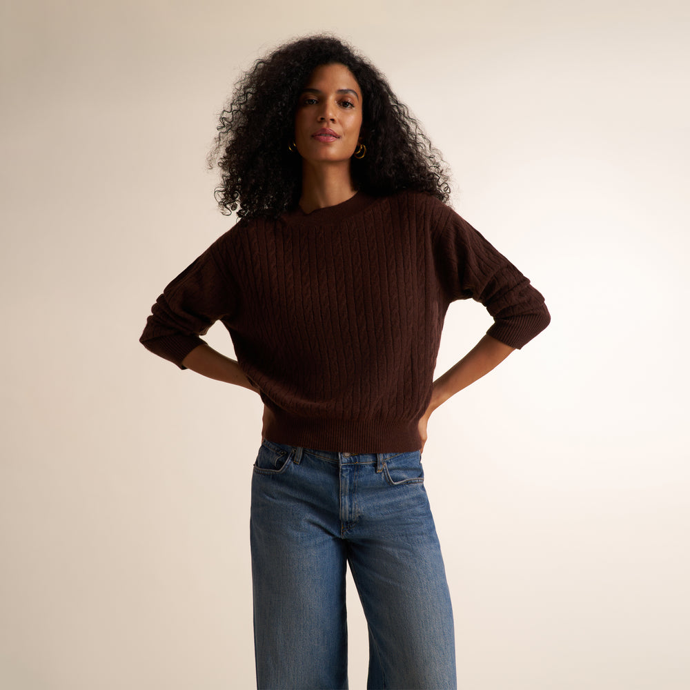 A woman with curly black hair wearing a dark brown cable-knit sweater and blue jeans, standing against a plain beige studio background.