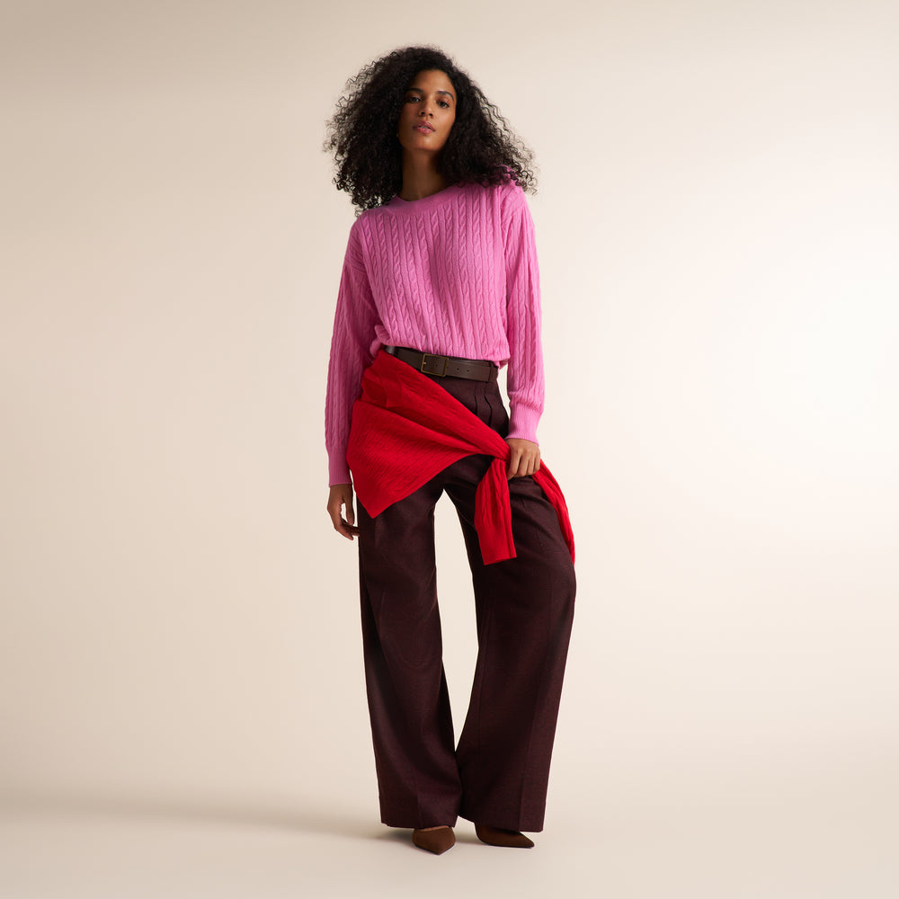 A model stands against a warm beige studio background wearing a bright pink cable-knit sweater, dark brown wide-leg trousers, and a vibrant red wrap tied around her waist.