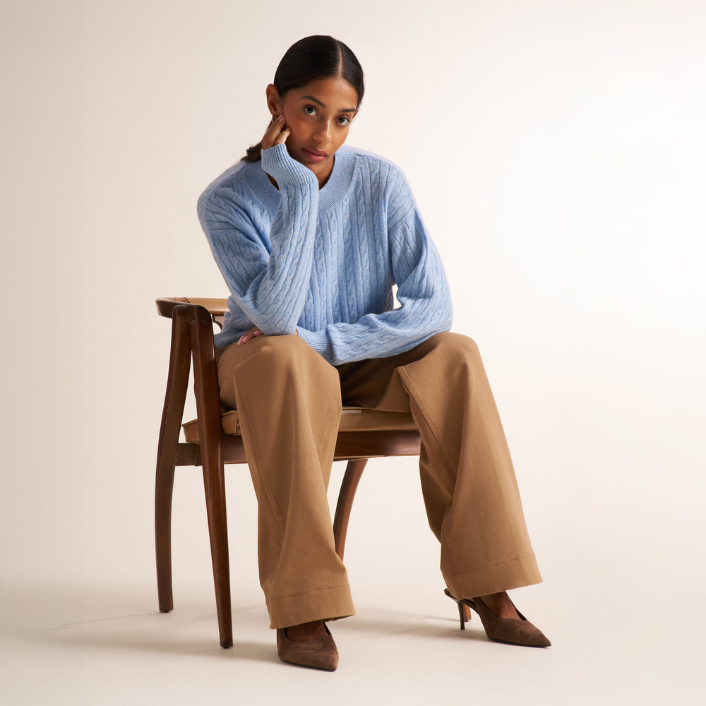 A brunette woman modeling a blue melange long-sleeve knit top with a a crew neck collar and cable knit detail, paired with saddle wide leg trousers, against a plain light beige studio background.