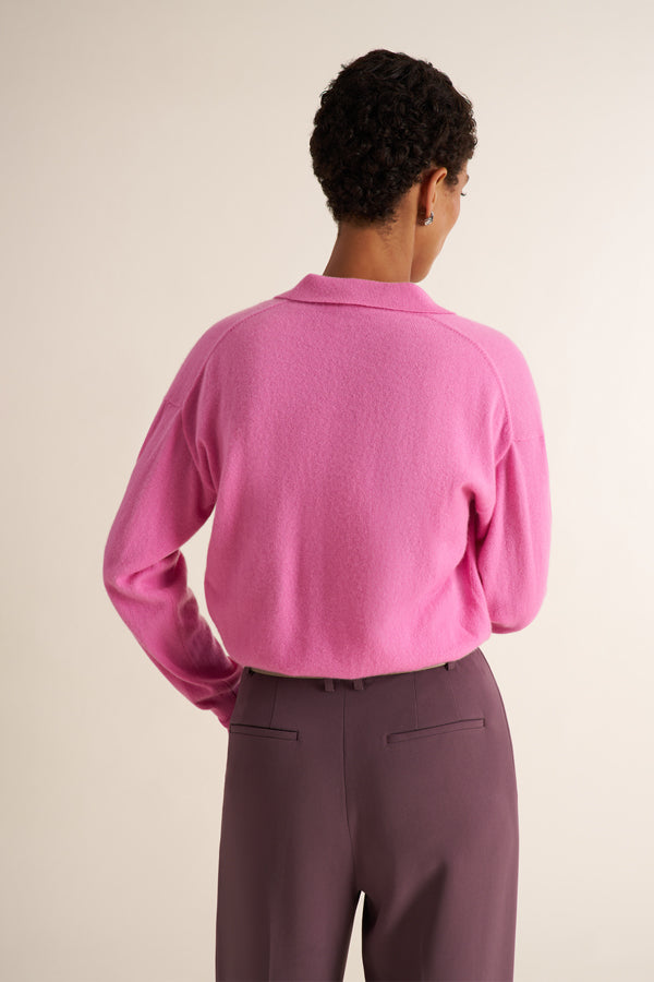 A brunette woman modeling the back of a pink long-sleeve knit top with a split V-neck collar, paired with purple trousers, against a plain light beige studio background.