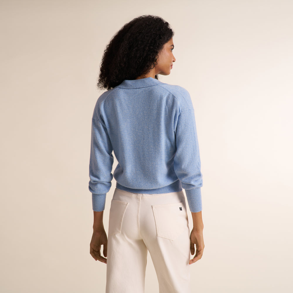 A curly brunette woman modeling a blue melange long-sleeve knit top with a split V-neck collar, paired with ivory wide leg trousers, against a plain light beige studio background.