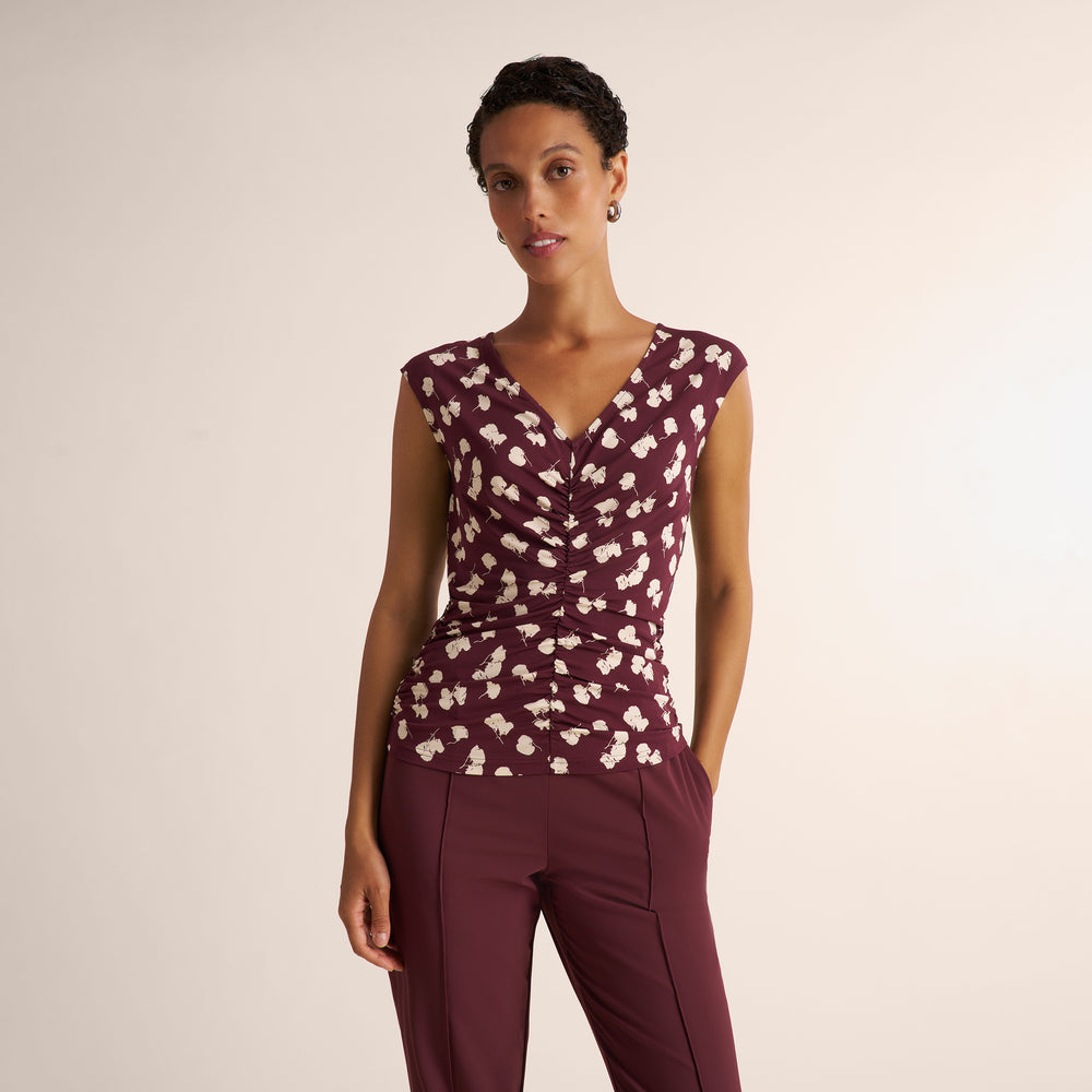 A woman modeling a sleeveless V-neck top in burgundy with a white floral pattern, paired with matching burgundy trousers, against a plain light beige studio background.