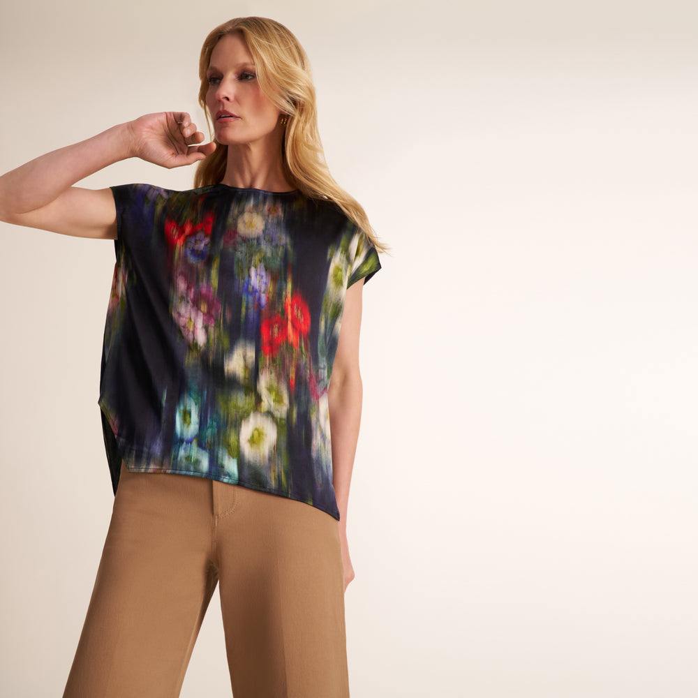 A woman modeling a short-sleeve top with a dark navy base and a colorful floral print, paired with tan trousers, against a plain light beige studio background.