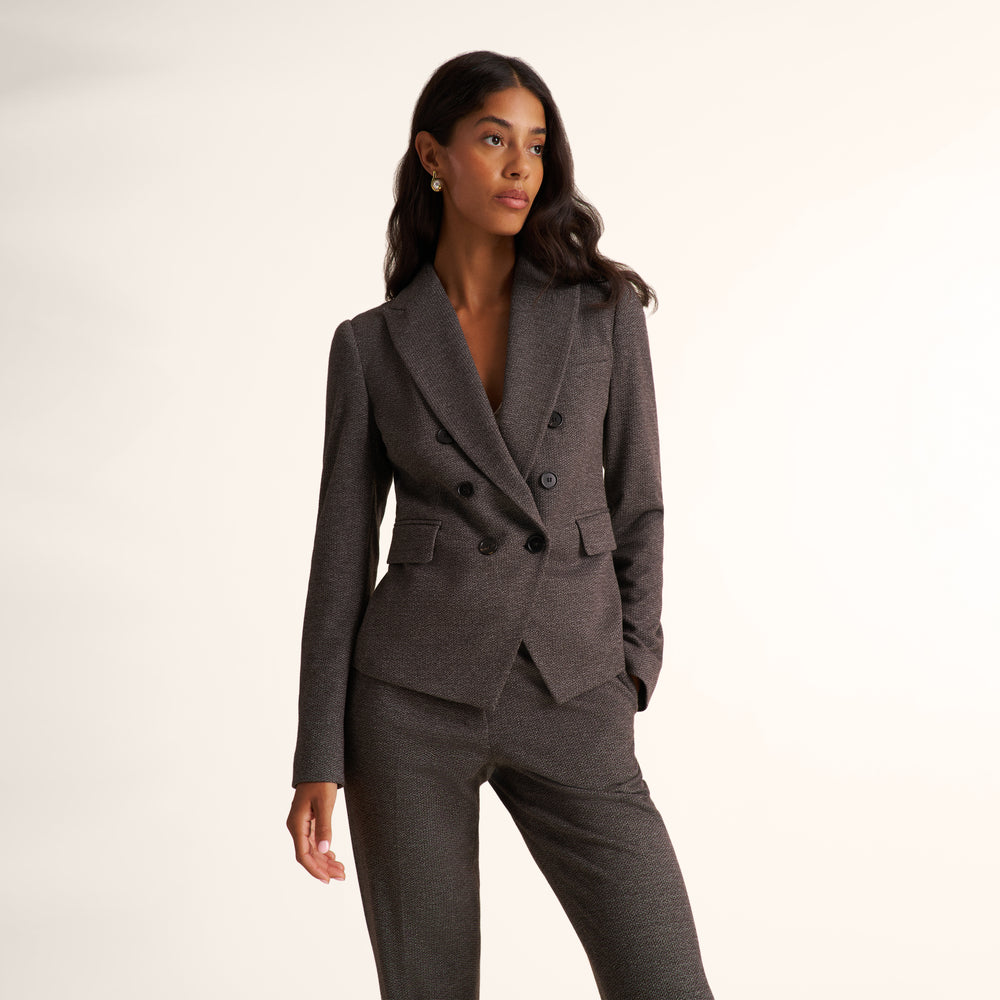 A woman modeling a dark gray double-breasted blazer with matching trousers against a clean, off-white studio background.