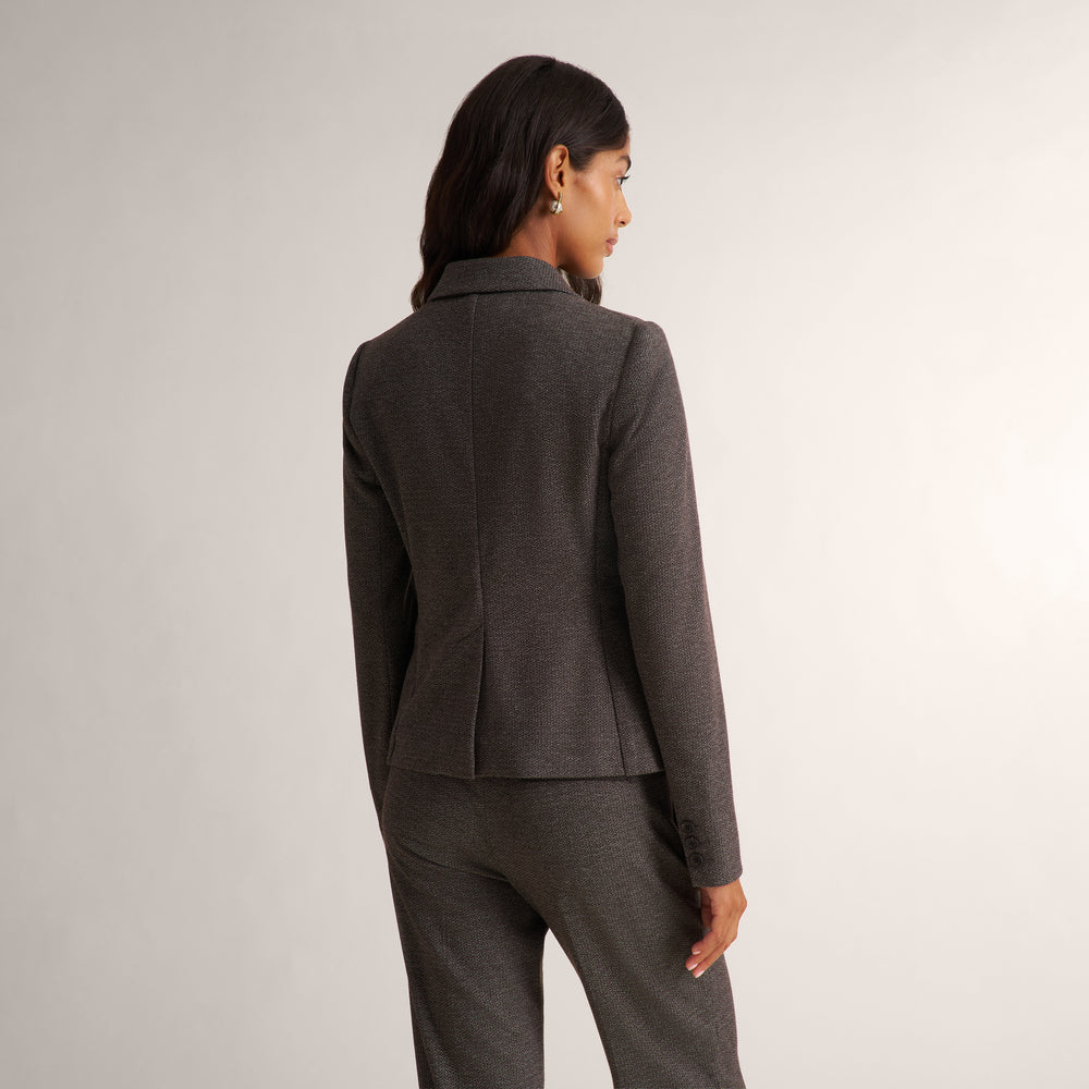 A woman shown from the back wearing a dark gray textured blazer and matching trousers against a light gray studio background.