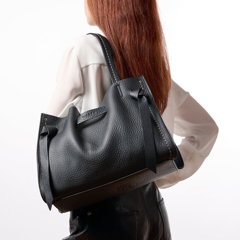 A woman shown from the back wearing a black pebbled leather shoulder bag slung over her shoulder, paired with a white blouse and black leather pants, against a plain white background.
