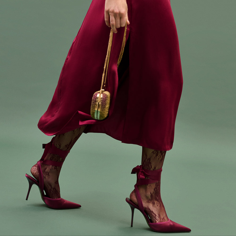 Person wearing a burgundy dress and high heels holding a gold handbag on a green background