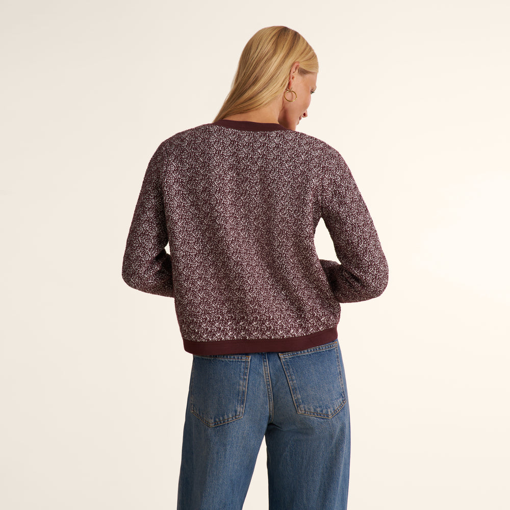 A woman viewed from the back wearing a textured, speckled knit sweater in a dark plum-brown color with solid ribbed trim, paired with blue jeans, against a plain off-white background.
