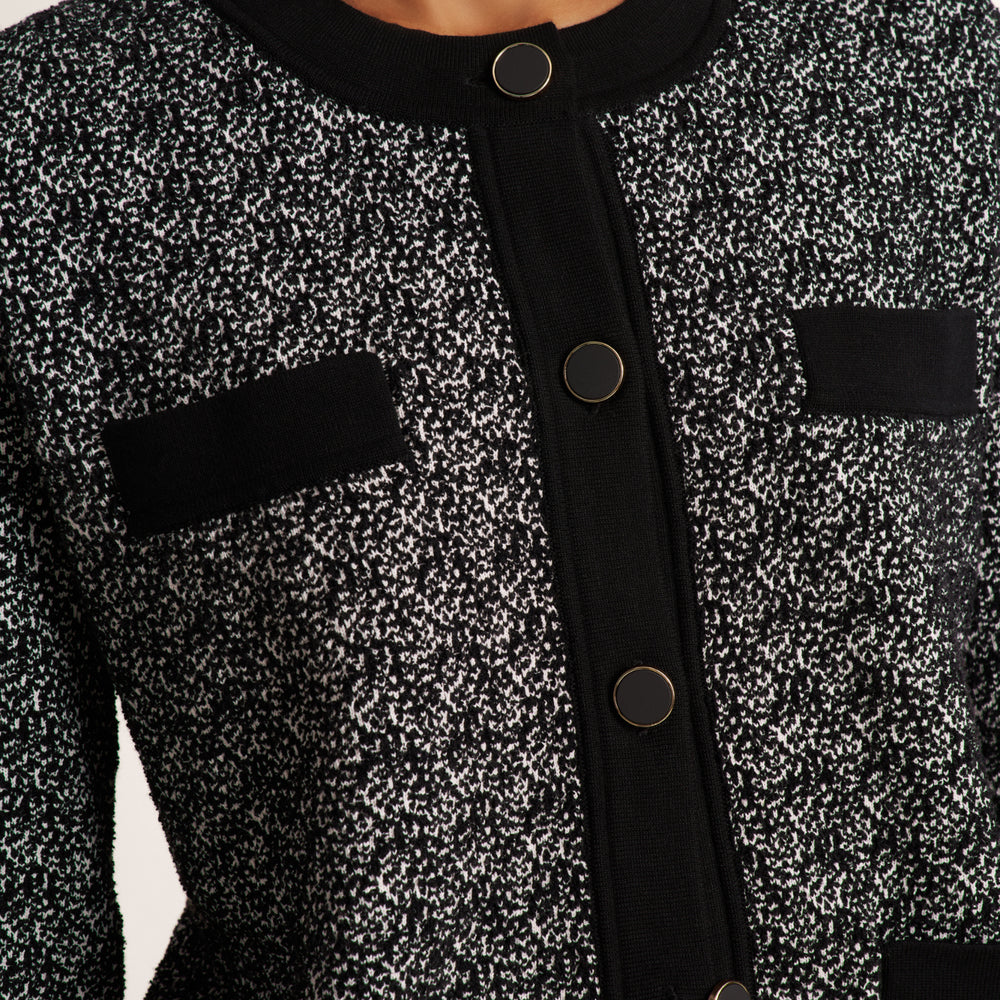Close-up of a black and white speckled tweed jacket with solid black trim, button closure, and flap pockets.
