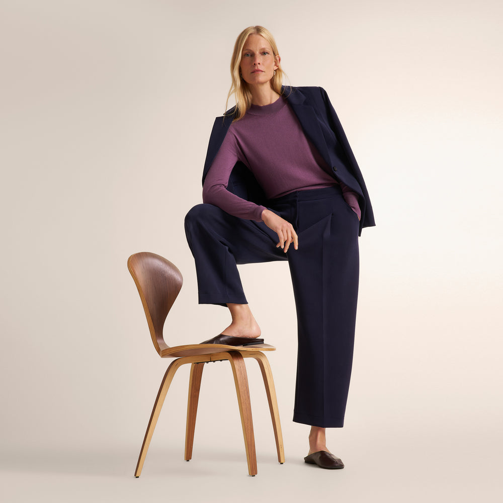 A woman models a dark navy blue two-piece suit consisting of a blazer and wide-leg trousers, layered over a purple long-sleeve top. She is posed sitting on a wooden chair against a solid light beige studio background.