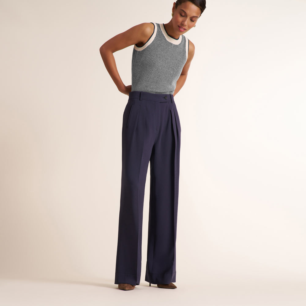 A model wearing high-waisted navy blue wide-leg trousers with pleats and a gray sleeveless knit top with a cream collar, standing against a solid off-white studio background.