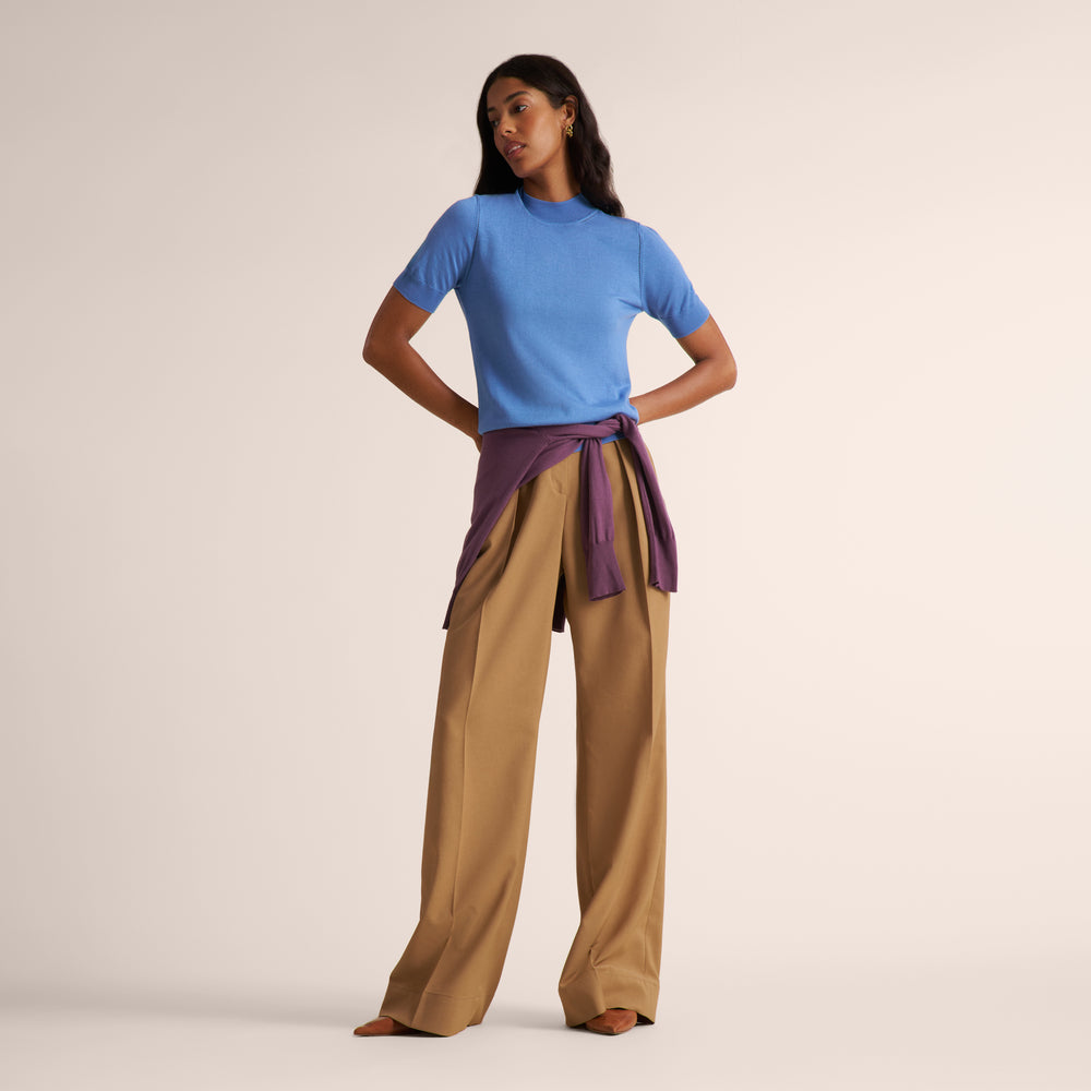 Woman wearing a mock neck short sleeve blue splendor sweater with wide leg beige pleated pants and purple sweater tied around her waist